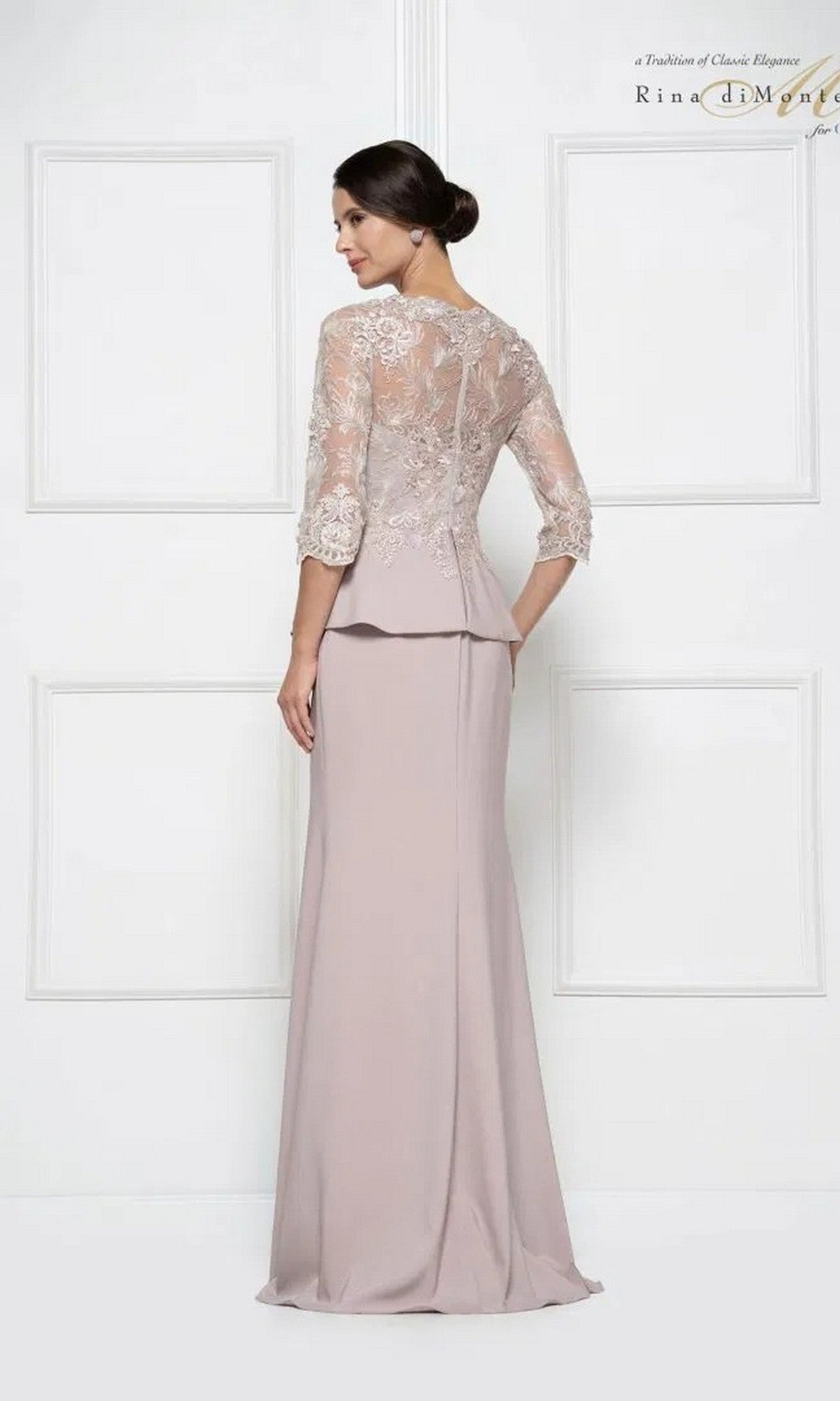 Woman in a Rina Di Montella RD2685 long formal dress featuring pale mauve color, embroidered lace sleeves, and a sheer lace back, standing sideways against a white paneled wall.